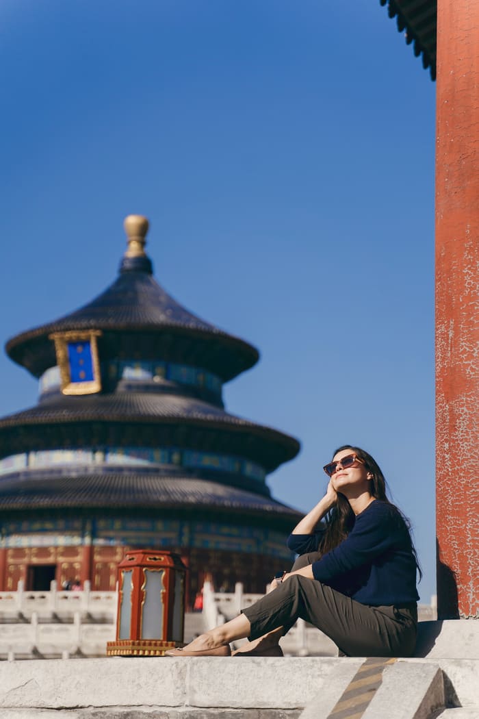 Una mujer joven con gafas de sol sentada tranquilamente frente al emblemático Templo del Cielo en Pekín, China, durante un día soleado de cielo despejado. La imagen captura una experiencia de turismo cultural y relajación en un sitio histórico. expertos en viajes a China en un templo tradicional