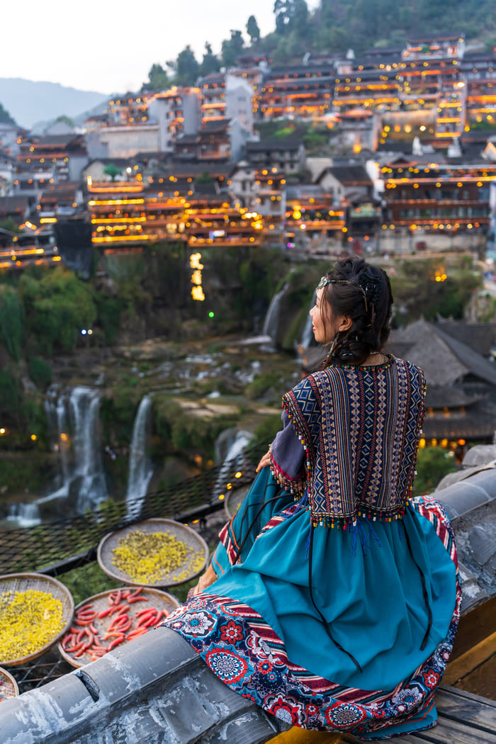 Mujer con vestimenta étnica tradicional china sentada de espaldas, observando la vista nocturna de una ciudad antigua iluminada junto a una cascada, con cestas de hierbas secas en primer plano. viajes personalizados a China
