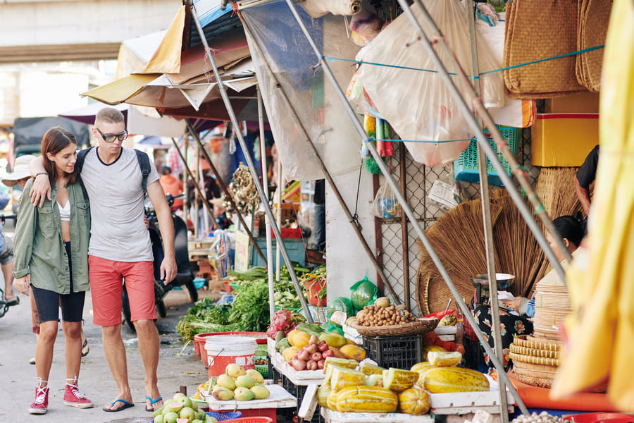 Pareja joven de turistas caminando abrazados por un mercado callejero tradicional en China, rodeados de puestos de frutas frescas y productos artesanales locales.