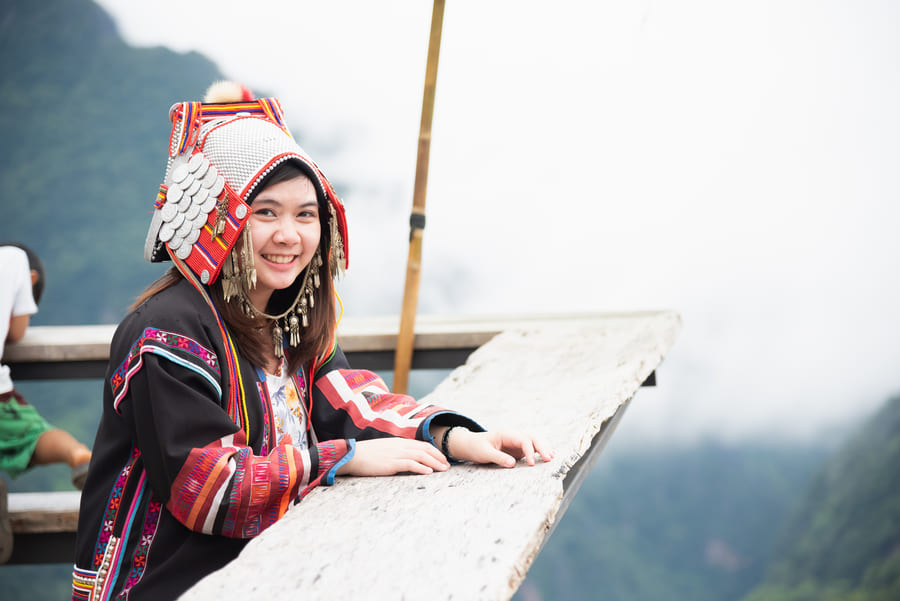 Mujer joven sonriente Itinerarios Personalizadosvistiendo el traje tradicional de la etnia Hmong, caracterizado por su intrincado tocado ornamental y bordados coloridos, apoyada en una plataforma de madera con un paisaje montañoso nublado de fondo.