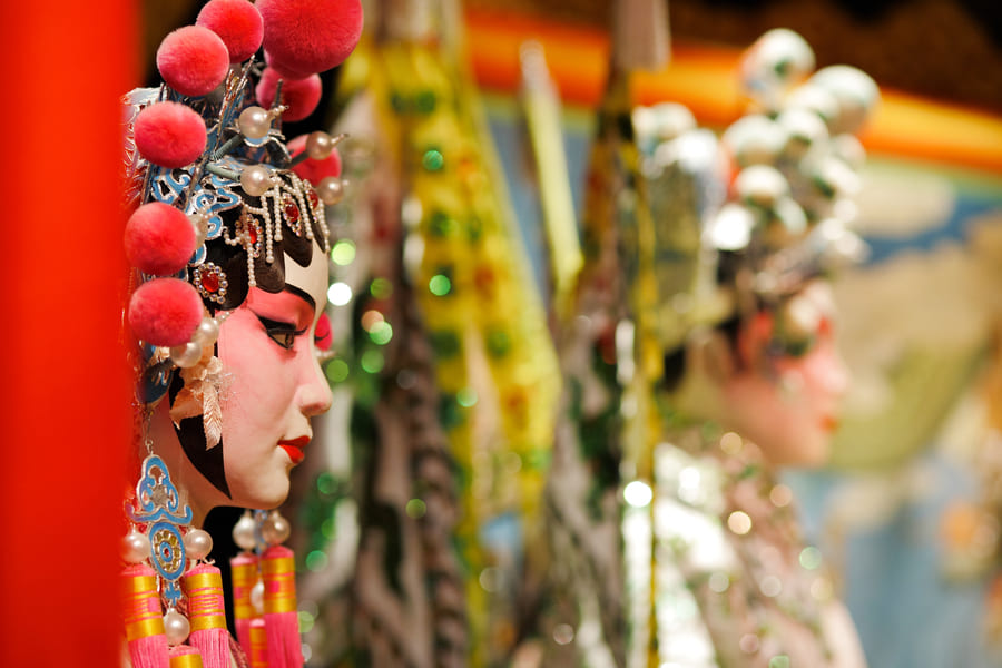 actriz de la Ópera de Pekín con maquillaje facial tradicional, tocado ornamentado con pompones rosas y pedrería, preparándose para una representación teatral en China. Itinerarios Personalizados