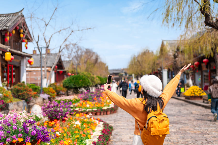 Una turista de espaldas, Itinerarios Personalizadosvistiendo un suéter naranja, gorro de invierno y mochila, tomando una fotografía con su celular en las calles de la ciudad antigua de Lijiang, China, rodeada de coloridos jardines de flores y arquitectura tradicional.
